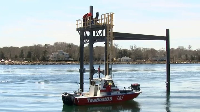 A red and white boat sits on the water next to a structure known as the Bourne Tidal Testing Site. A man stands on the structure above the boat while others look up at him.